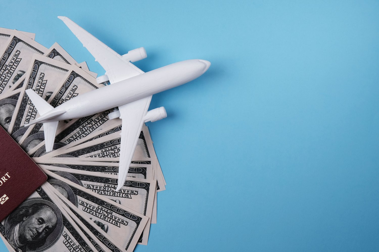 A model airplane rests on top of a fan of U.S. hundred-dollar bills and a passport, set against a blue background—symbolizing the financial aspects of aviation and international travel.