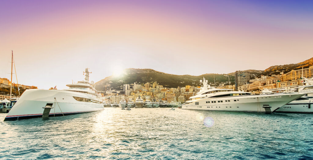 Private Jet to Monaco Yacht Show 2025 – Routes, Aircraft & Costs 2 Luxury superyachts docked in Port Hercule at sunset during the Monaco Yacht Show, with the city skyline and mountains in the background.