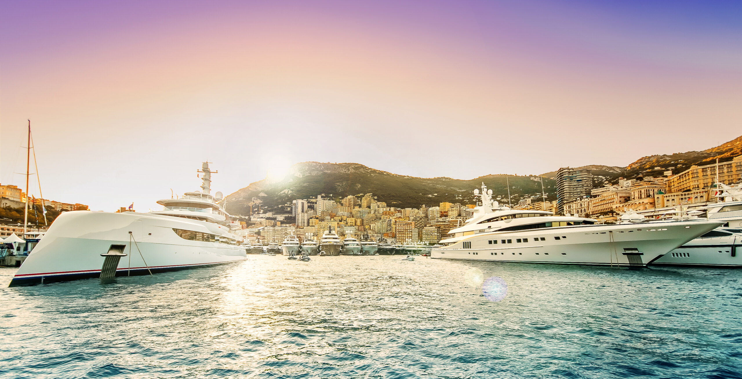 Luxury superyachts docked in Port Hercule at sunset during the Monaco Yacht Show, with the city skyline and mountains in the background.